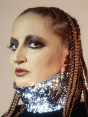 Futuristic Glam - Singer Mina with braided hair and dramatic makeup wears a metallic, reflective collar. The makeup features bold, smoky eyeshadow and glossy lips. The background is a soft beige, and the person gazes slightly to the side.