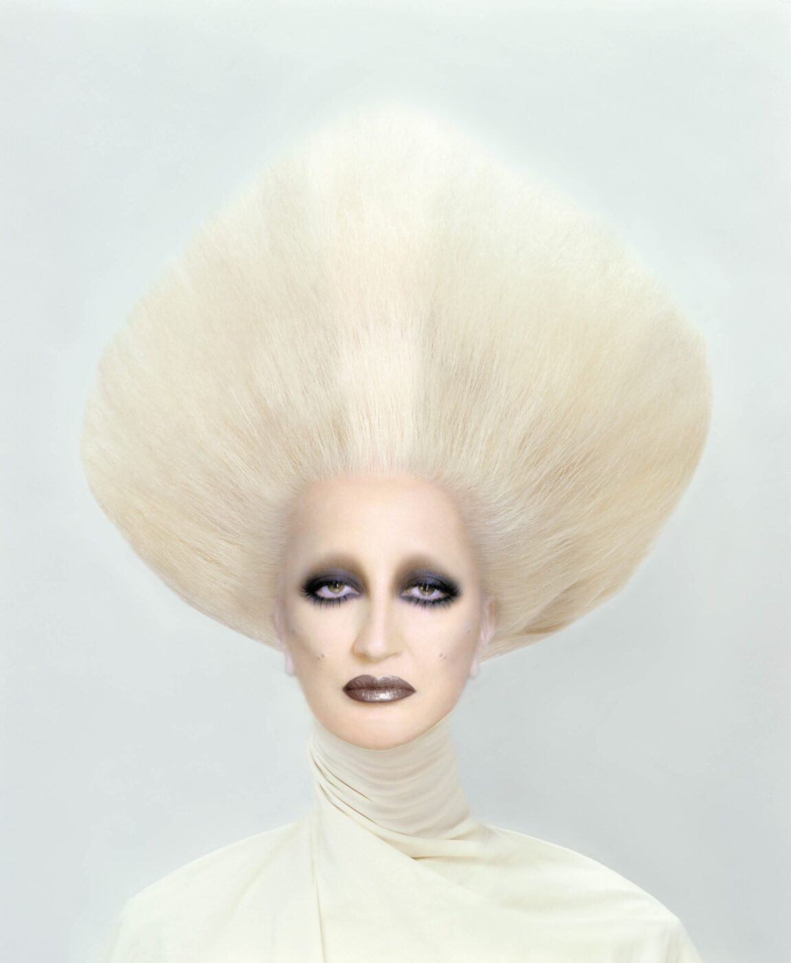 Mina on the cover of Veleno with a dramatic, voluminous, white hairstyle styled into a large triangular shape. They have a pale complexion, dark eye makeup, and dark lipstick. They are wearing a light, high-collared garment against a plain background.