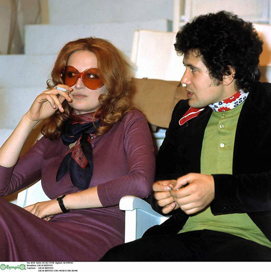 Mina, with long red hair and large orange sunglasses sits in a white chair, smoking. She wears a purple dress and a patterned scarf. Lucio Battisti, with curly hair, wearing a black jacket and green shirt, sits beside her, holding a cigarette.