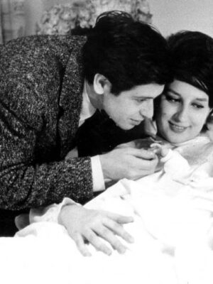 Mina and Corrado Pani lovingly looks at their newborn baby, who is resting on a hospital bed. The father, wearing a suit, gently touches the babys hand, while the mother, with styled hair, smiles warmly. The room is softly lit, with floral curtains in the background.