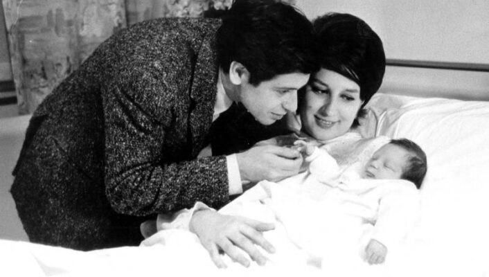 Mina and Corrado Pani lovingly looks at their newborn baby, who is resting on a hospital bed. The father, wearing a suit, gently touches the babys hand, while the mother, with styled hair, smiles warmly. The room is softly lit, with floral curtains in the background.