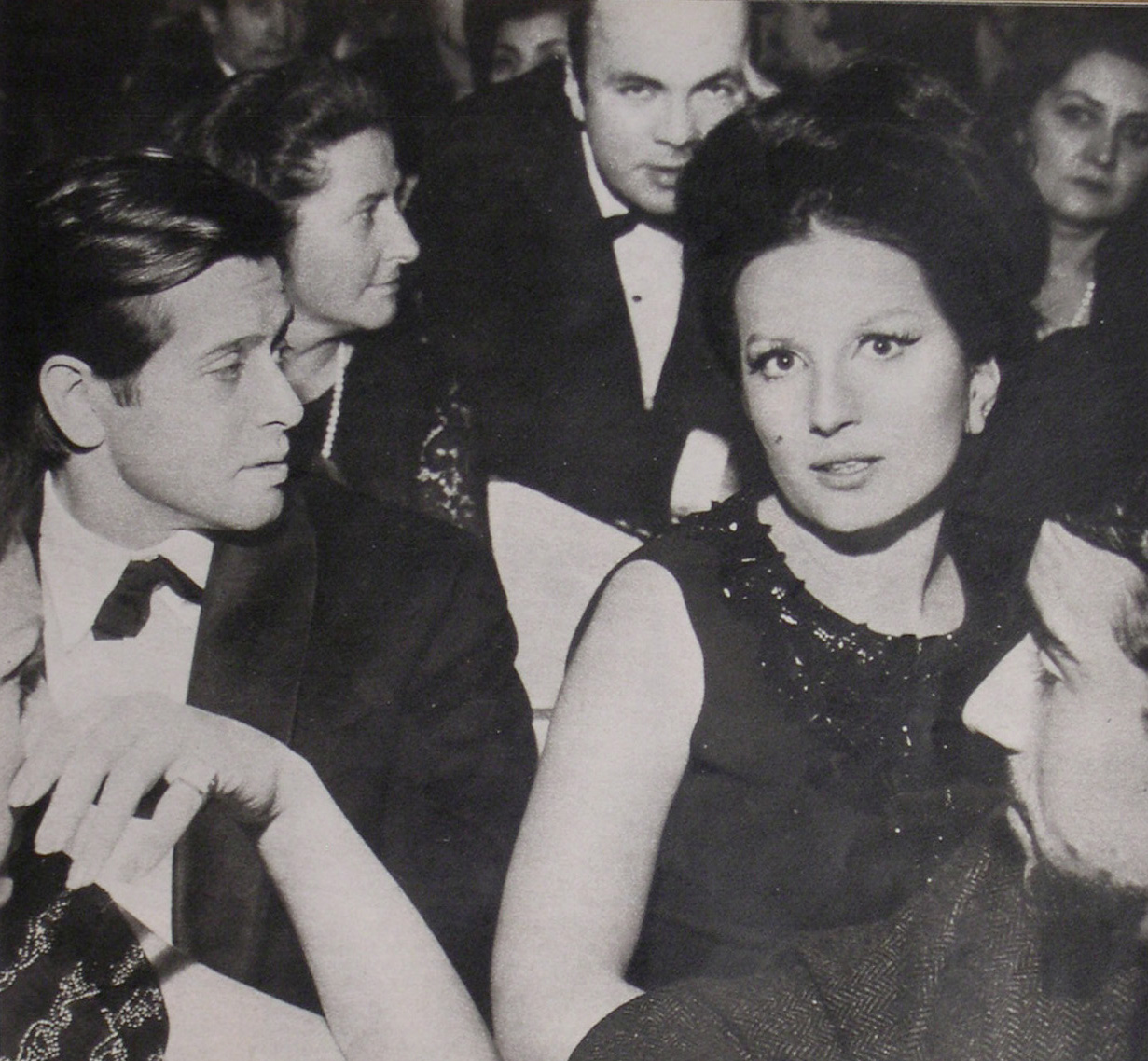 Black and white image of a formal event. Mina in a sleeveless dress sits next to Corrado Pani in a suit and bow tie. They are surrounded by other people in formal attire, some looking at the camera and others looking in different directions.
