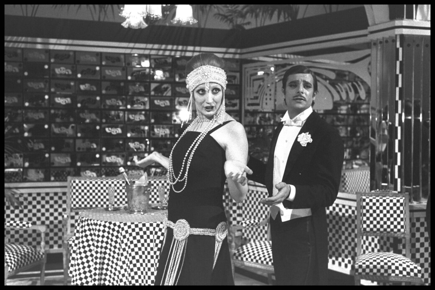 Mina and Giancarlo Giannin - Two people dressed in vintage 1920s attire appear in a retro-themed setting. The person on the left wears a beaded headpiece and a flapper dress, while the person on the right dons a tuxedo and bow tie. The background features checkered decor.