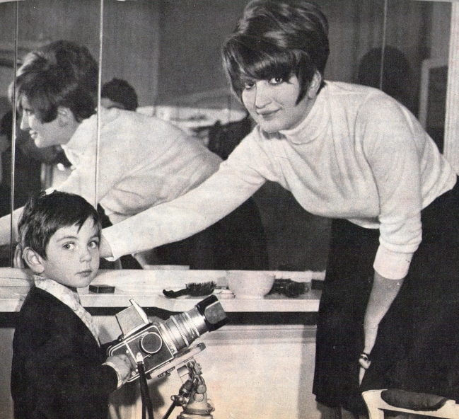 Mina Mazzini with short hair stands and slightly bends toward a young boy who holds an old-fashioned camera on a tripod. Theres a mirror behind them reflecting her image. The boy is looking at the camera. The setting appears to be indoors.