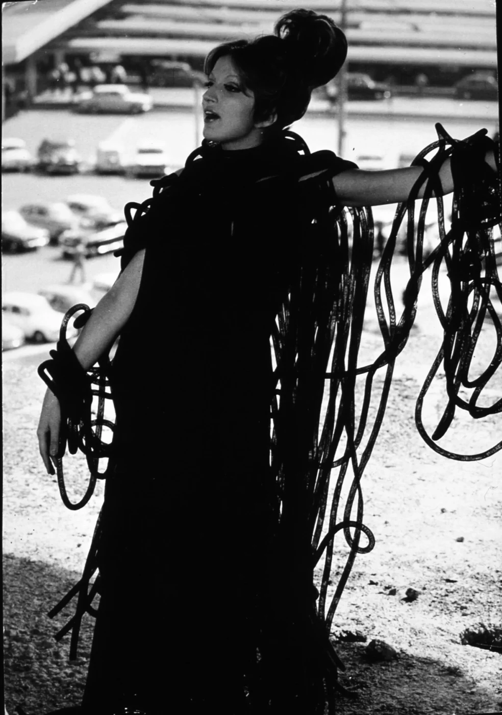 Mina wearing a long, flowing black dress with numerous ribbon-like extensions poses dramatically outdoors. The background features parked cars and a bridge structure. The image is in black and white, emphasizing contrast and texture.