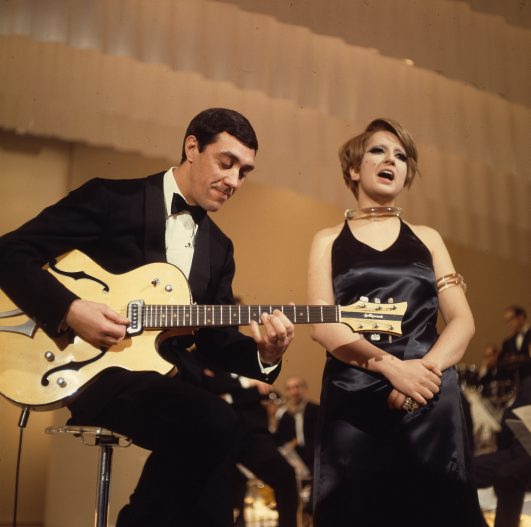 Performing Duo - Un uomo in smoking nero suona una chitarra elettrica seduto su uno sgabello. Una donna in abito nero senza maniche canta accanto a lui. Entrambi sono su un palco con uno sfondo sfocato di musicisti seduti.