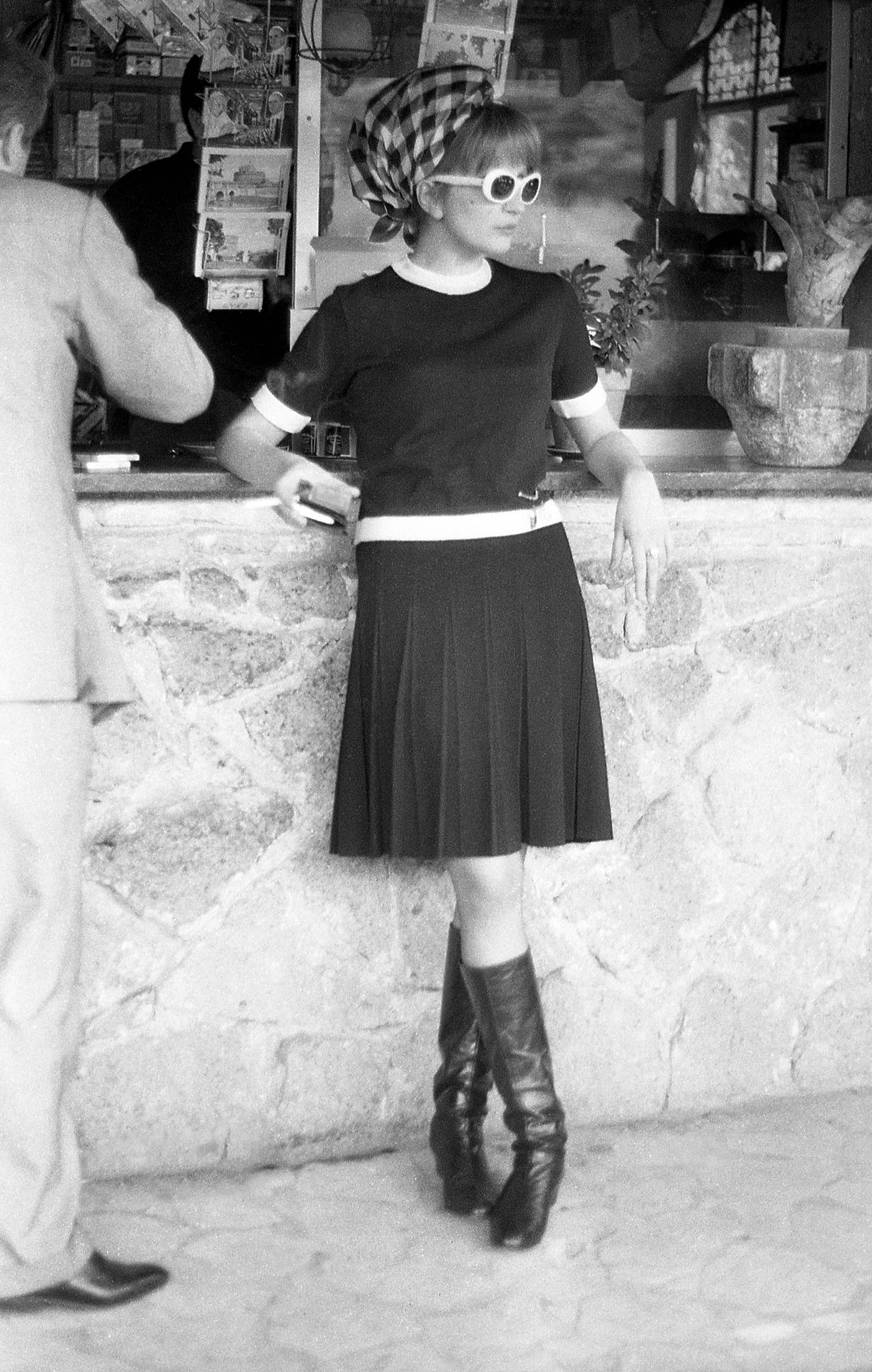 Black and white photo of Mina in a dark dress and knee-high boots, leaning against a stone wall. She wears sunglasses and a patterned headscarf. A person in a suit is partially visible on the left, near a kiosk displaying postcards.