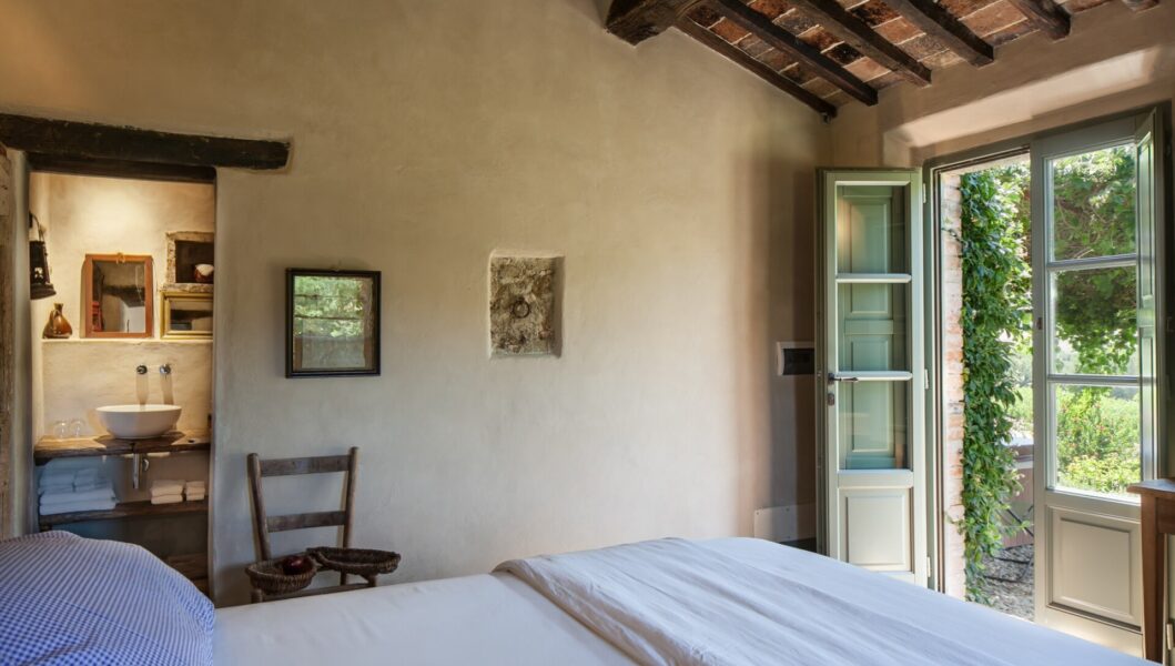 Cozy Bedroom Escape - Accogliente camera da letto con letto matrimoniale, soffitto rustico in legno e una porta che conduce a un rigoglioso giardino. La stanza dispone di un piccolo bagno con lavandino e specchio, una sedia in legno e una finestra aperta con verde allesterno.