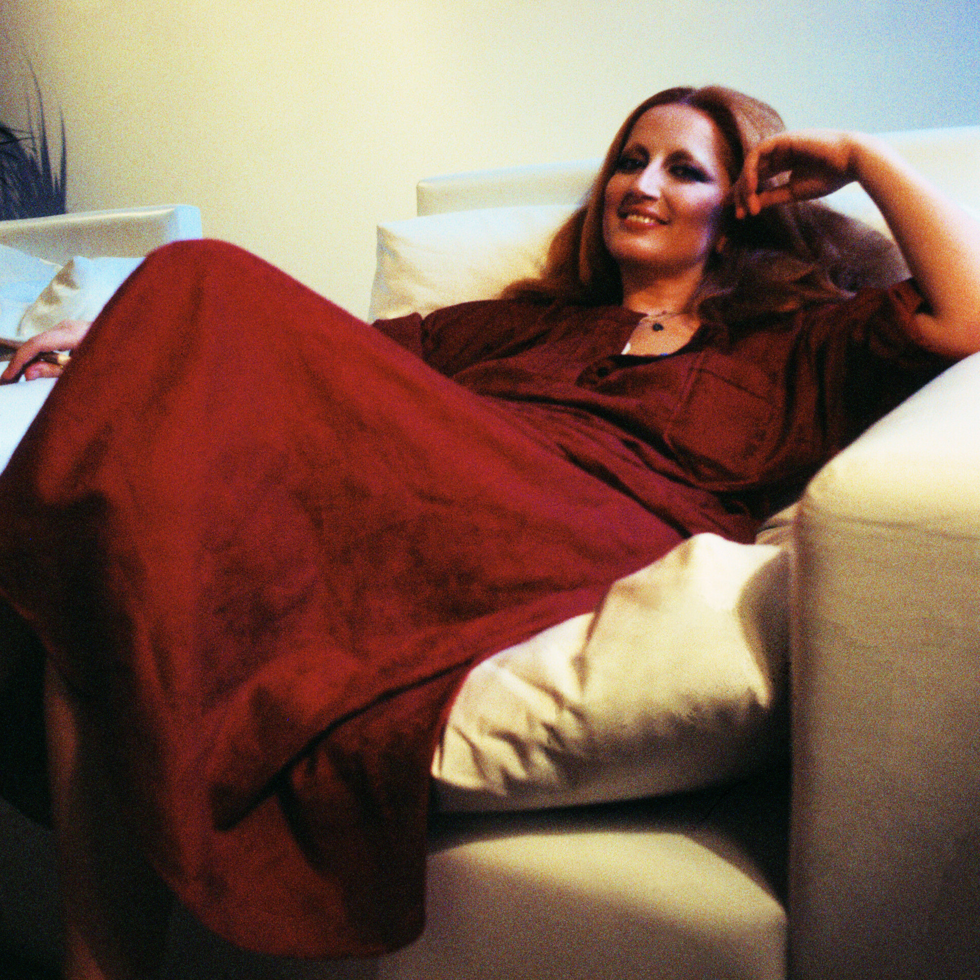 Mina Mazzini with long hair wearing a red dress, lounging comfortably on a white sofa. They have a relaxed expression, with one arm resting on the back of the sofa and a leg tucked under the other. The setting appears to be indoors.