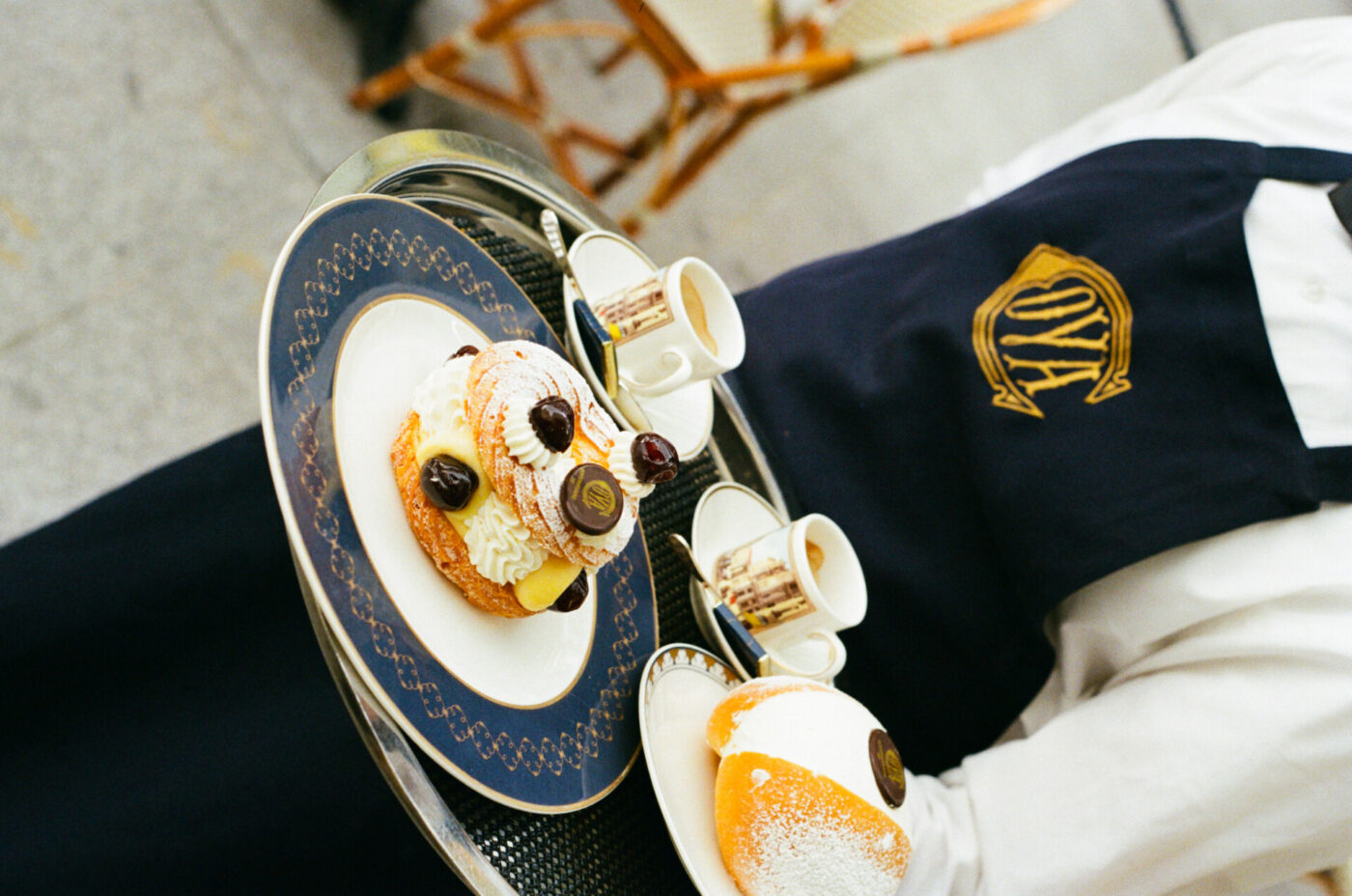 The Sweet Indulgence" - Una persona che indossa un grembiule scuro regge un vassoio con un dessert guarnito con ciliegie e zucchero a velo, insieme a due piccole tazzine di espresso su piattini. L'ambientazione sembra essere un bar all'aperto con sedie di vimini.