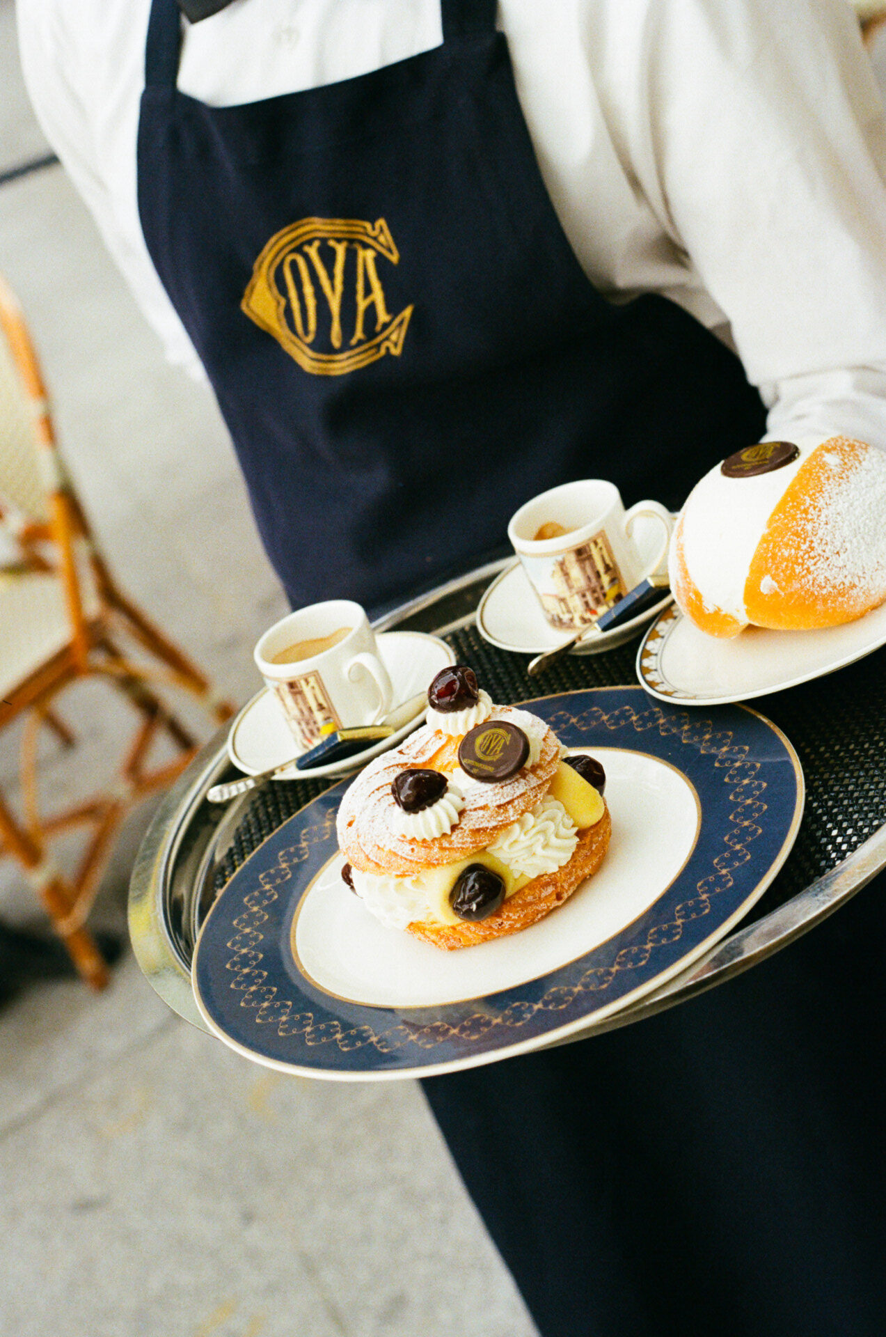 The Sweet Indulgence" - Una persona che indossa un grembiule scuro regge un vassoio con un dessert guarnito con ciliegie e zucchero a velo, insieme a due piccole tazzine di espresso su piattini. L'ambientazione sembra essere un bar all'aperto con sedie di vimini.