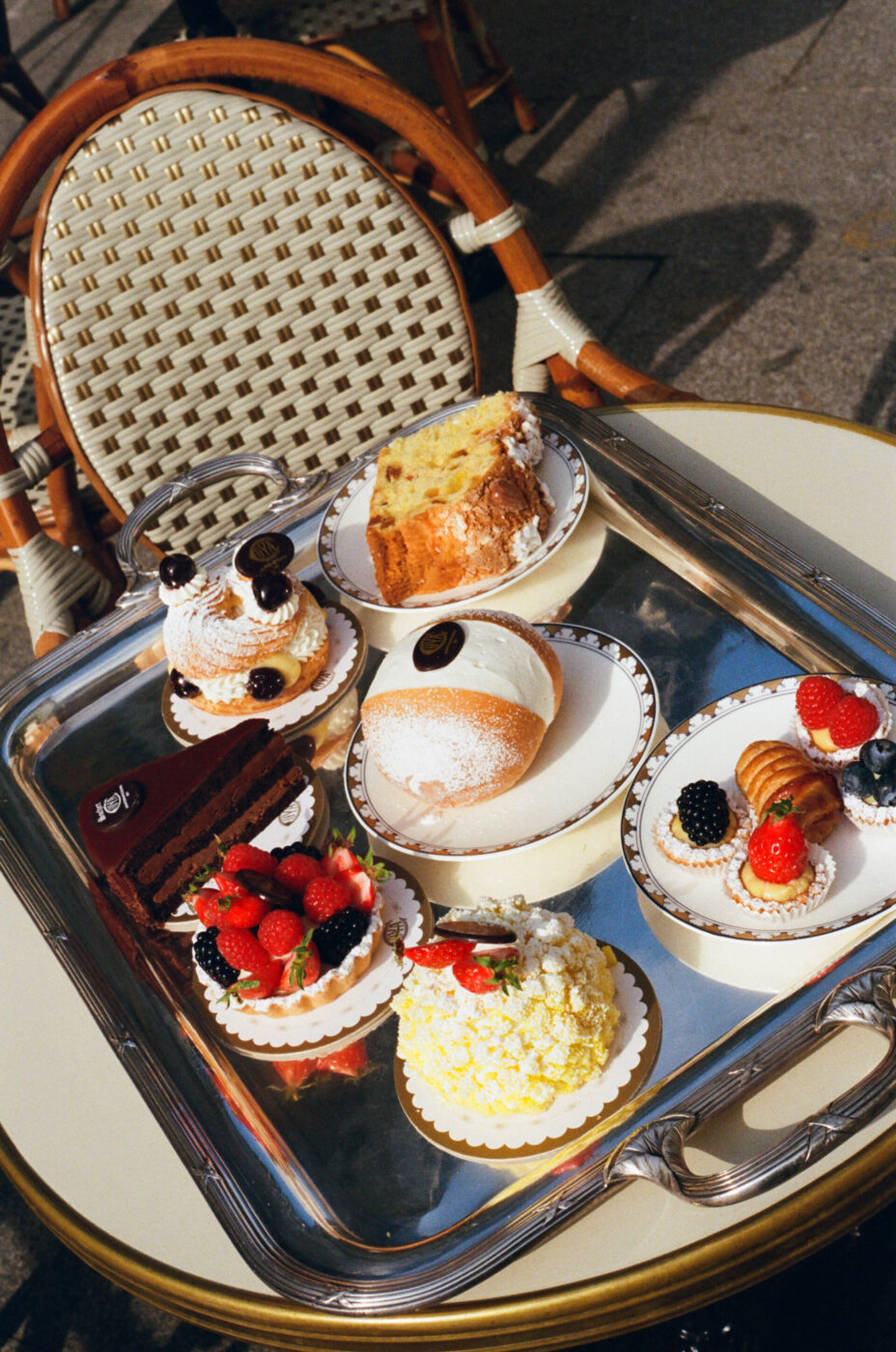 A Day at the Café" - Una varietà di pasticcini su un vassoio d'argento su un tavolo all'aperto. In primo piano ci sono crostate di frutta, un dessert al cioccolato, un pasticcino ripieno di crema e una fetta di torta. Una sedia intrecciata è sullo sfondo. La scena suggerisce una giornata di sole in un bar.