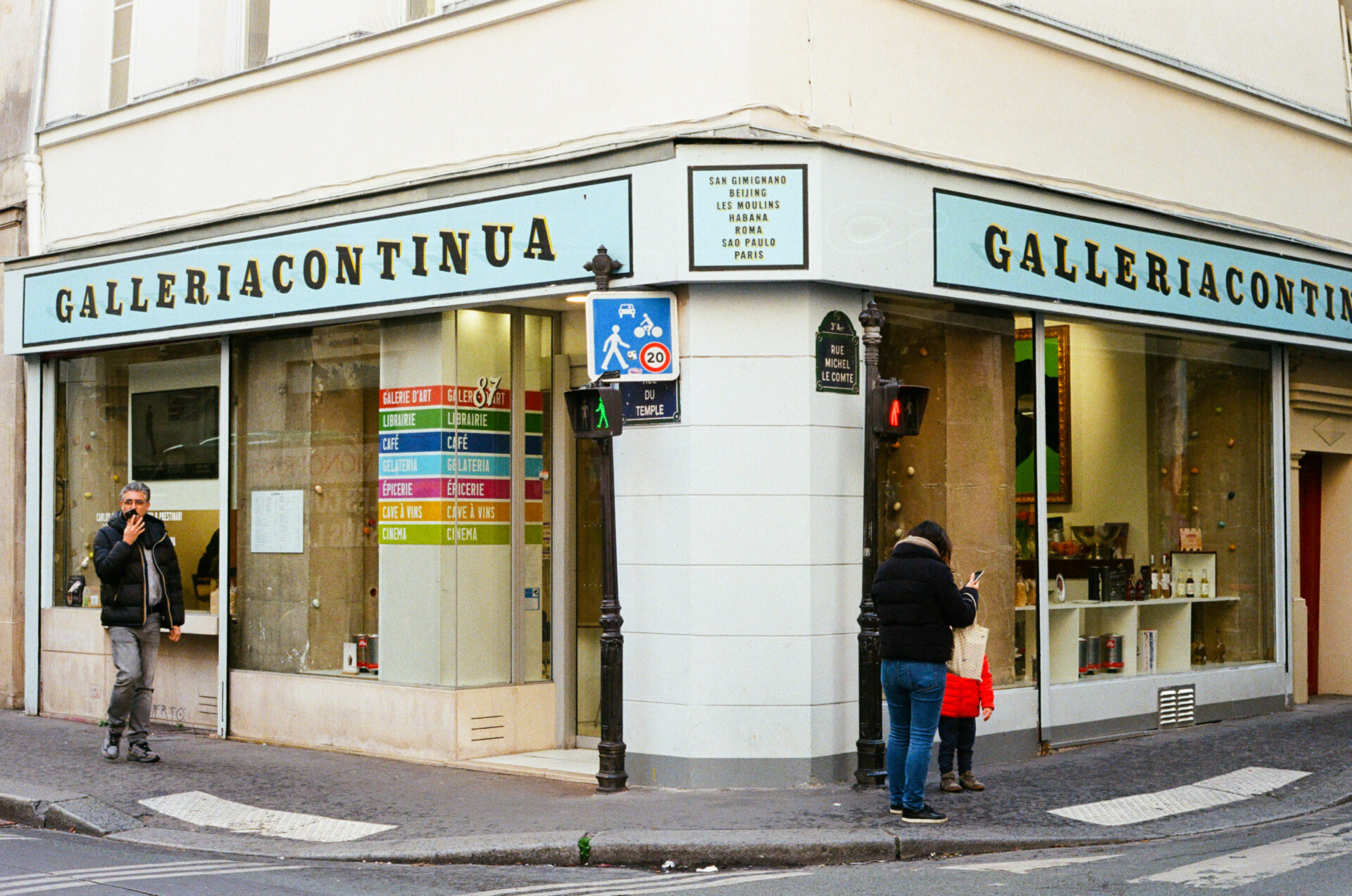 Street Corner Gallery" - Angolo di strada con una galleria d'arte moderna con vetrate. Due persone sono in piedi fuori, una usa un telefono e l'altra guarda l'arte attraverso la finestra. Un bambino con loro guarda dentro. Sono visibili vari cartelli e un semaforo.