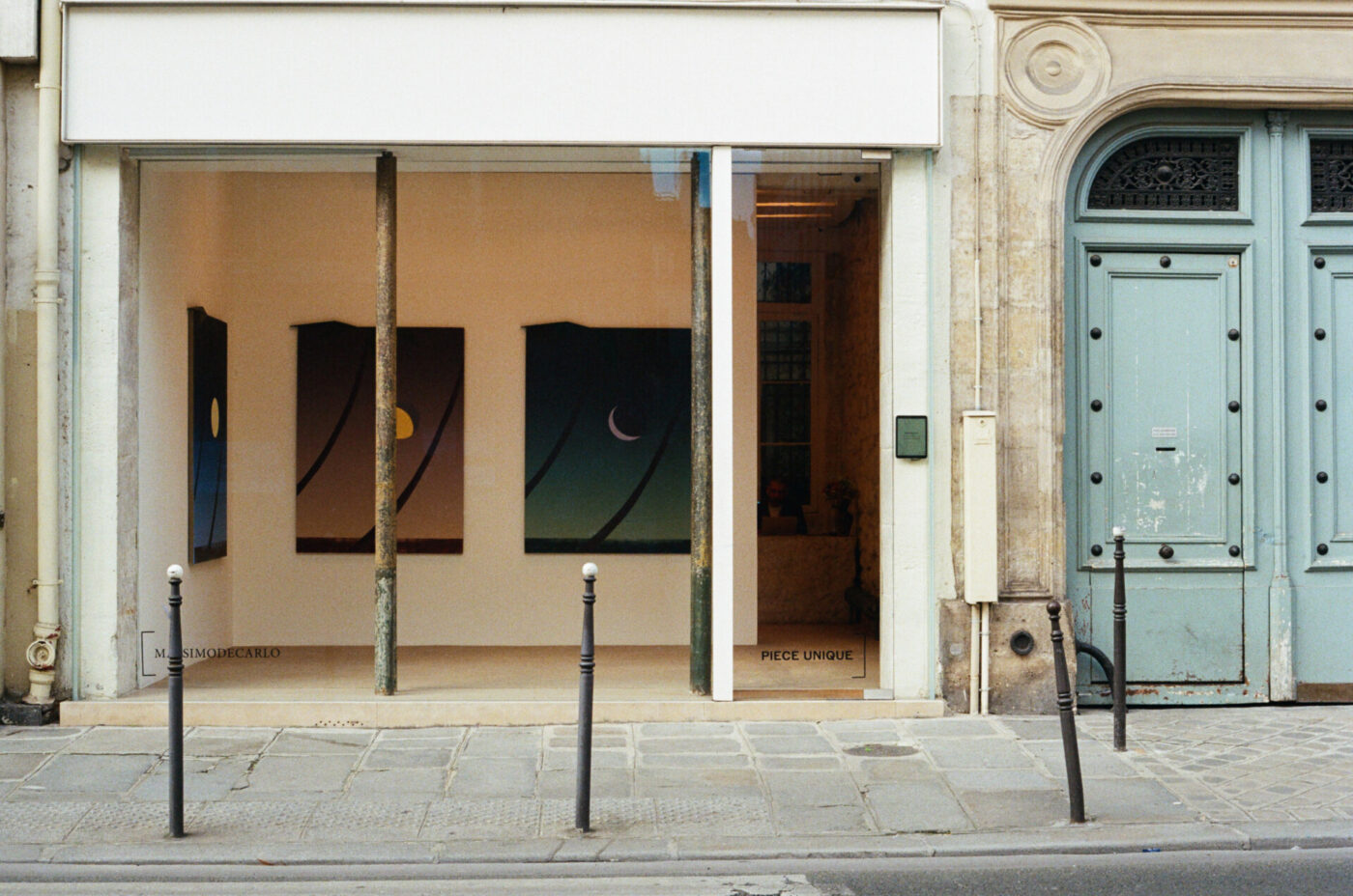 Mostra d'arte celeste - Una piccola galleria d'arte con una vetrina in vetro espone tre dipinti astratti con temi celesti. L'esterno dell'edificio è di colore chiaro, con una grande porta a due ante color verde acqua sulla destra e due sottili pali neri sul marciapiede di fronte.