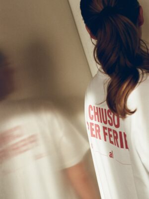 Chiuso Per Ferie Reflection - A person with long brown hair in a ponytail, wearing an Italy Segreta Chiuso Per Ferie T-Shirt, stands with their back to the camera. Their reflection appears on a shiny surface to the left.