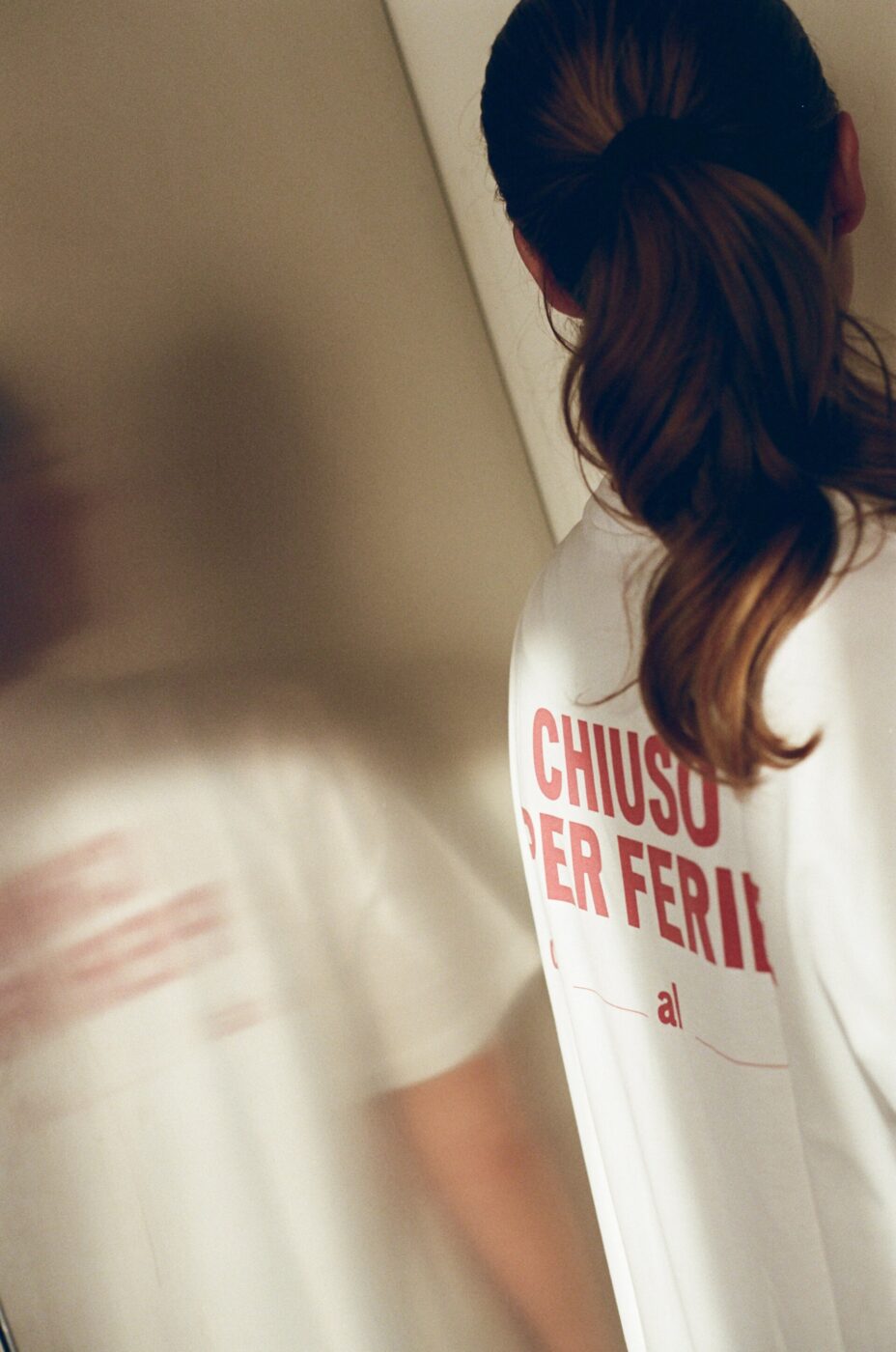 Chiuso Per Ferie Reflection - A person with long brown hair in a ponytail, wearing an Italy Segreta Chiuso Per Ferie T-Shirt, stands with their back to the camera. Their reflection appears on a shiny surface to the left.
