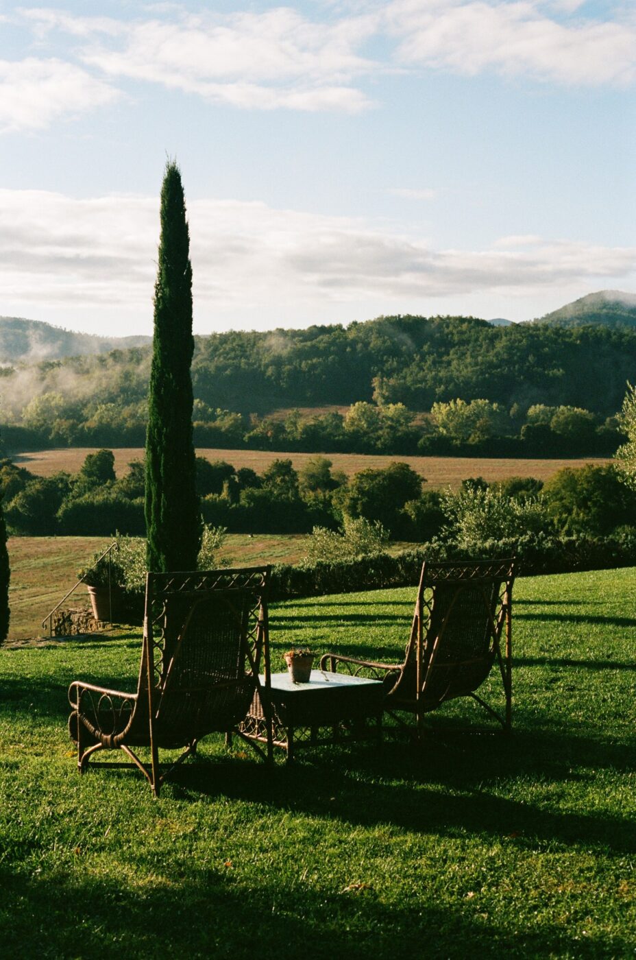 Borgo Santo Pietro - Two wrought iron chairs and a small table on a grassy lawn overlook a scenic landscape with rolling hills and a tall slender tree. Sunlight casts long shadows and clouds dot the blue sky, creating a peaceful atmosphere.