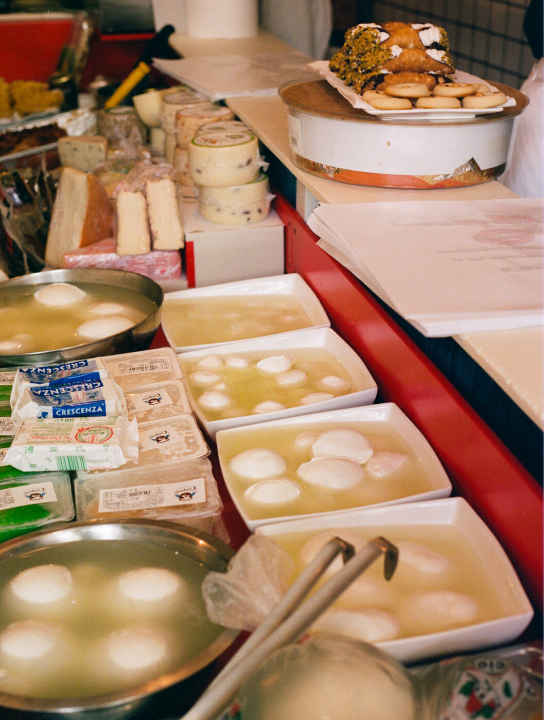 Mmmozza...! - A display of various fresh cheeses, including several bowls of mozzarella balls in water, blocks of other cheeses, and packaged cheese. Tongs lie in front, and a shelf above holds pastries with nuts.