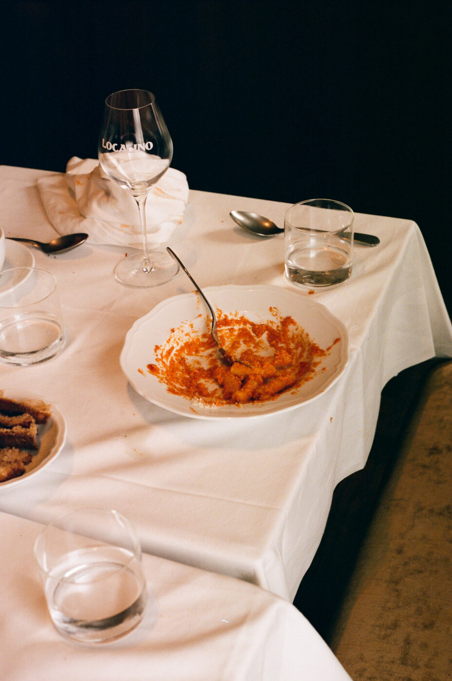 Piatto vuoto dopo pasto" - Un tavolo da pranzo vuoto con una tovaglia bianca regge un piatto con resti di un piatto di pasta al sugo rosso. Una forchetta è appoggiata sul piatto. Ci sono quattro bicchieri vuoti, un bicchiere con la scritta "Locarno" e tovaglioli usati sparsi in giro.