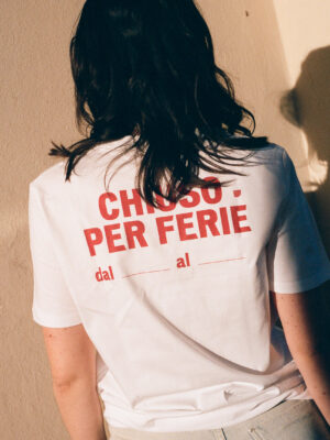 Chiuso Per Ferie T-Shirt - A person with long dark hair, wearing an Italy Segreta Chiuso Per Ferie T-Shirt featuring red text and date blanks, faces a wall. Sunlight casts a gentle shadow on the wall.