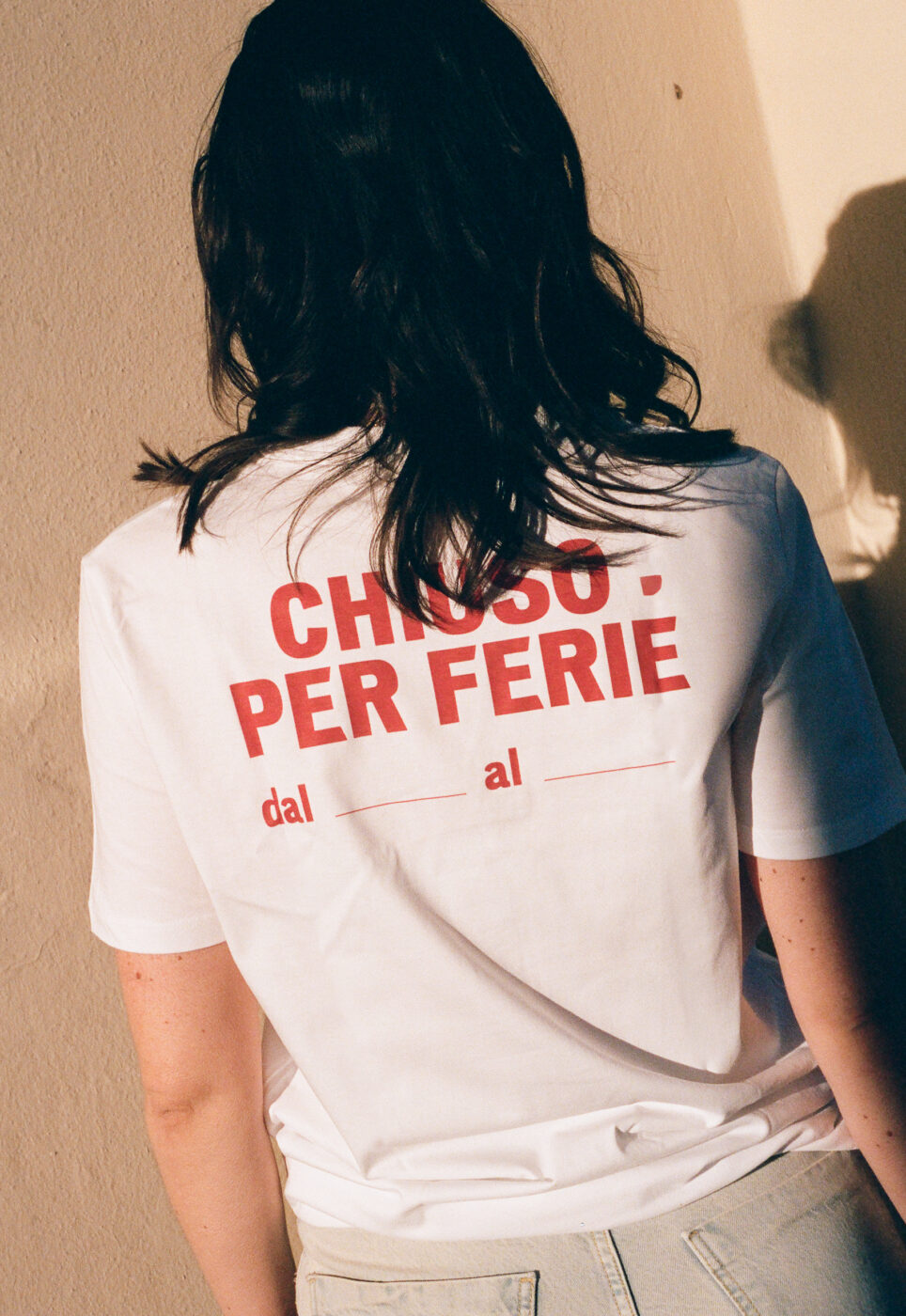 Chiuso Per Ferie T-Shirt - A person with long dark hair, wearing an Italy Segreta Chiuso Per Ferie T-Shirt featuring red text and date blanks, faces a wall. Sunlight casts a gentle shadow on the wall.