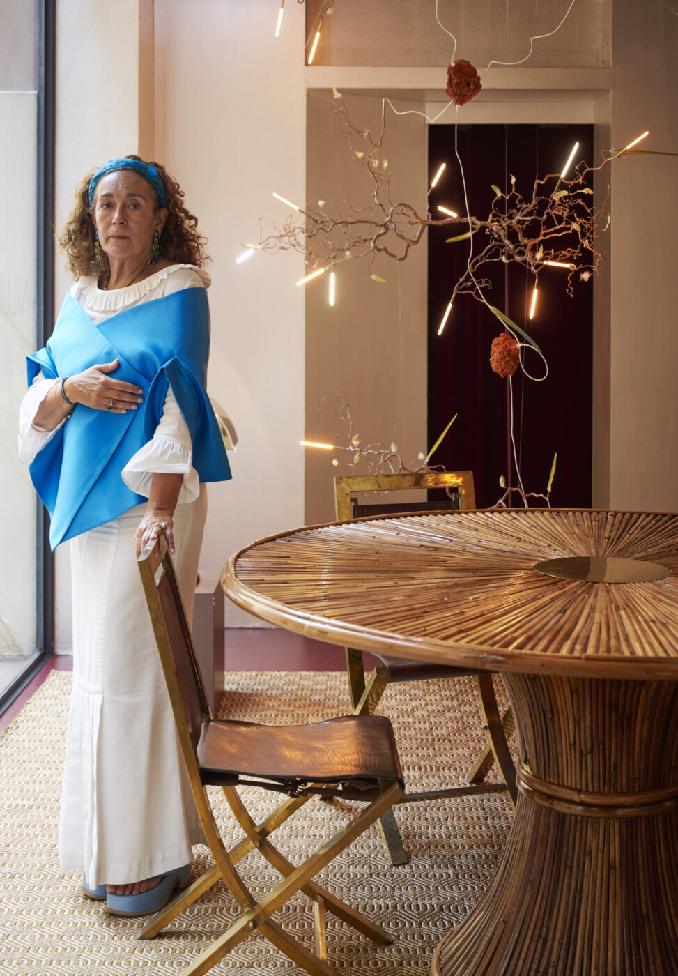 Nina Yashar; - A woman with curly hair stands indoors, wearing a blue shawl over a white dress. She is next to a bamboo table and chairs. The room is decorated with branches, orange pom-poms, and hanging lights. She gazes out of a large window to her right.