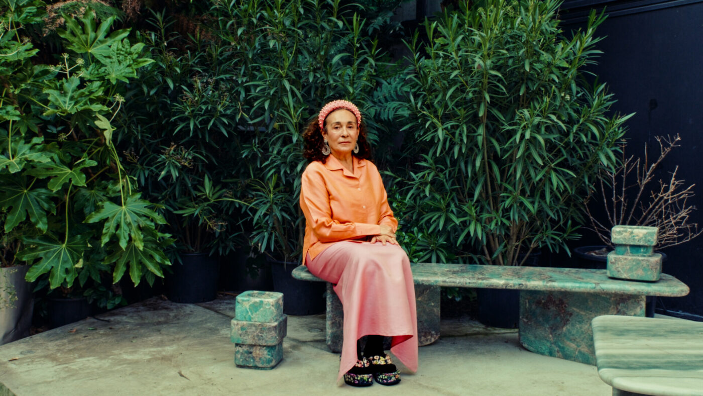 Nina Yashar wearing an orange shirt, pink skirt, and pink headscarf sits on a green stone bench surrounded by lush green plants. The scene is tranquil and set in an outdoor garden area.
