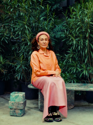 Nina Yashar wearing an orange shirt, pink skirt, and pink headscarf sits on a green stone bench surrounded by lush green plants. The scene is tranquil and set in an outdoor garden area.