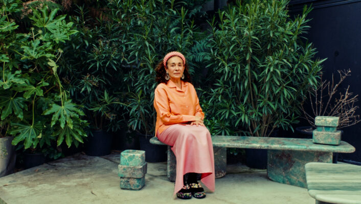 Nina Yashar wearing an orange shirt, pink skirt, and pink headscarf sits on a green stone bench surrounded by lush green plants. The scene is tranquil and set in an outdoor garden area.