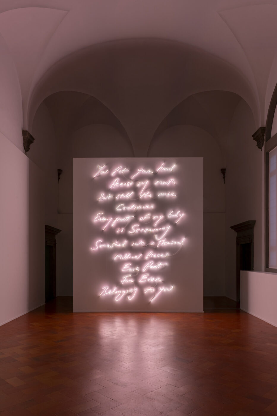 A Neon Evening - A dimly lit room with vaulted ceilings featuring a large neon art installation. The neon lights form cursive text in white on a square panel. The floor is polished wood, and tall windows are partially visible on the right.