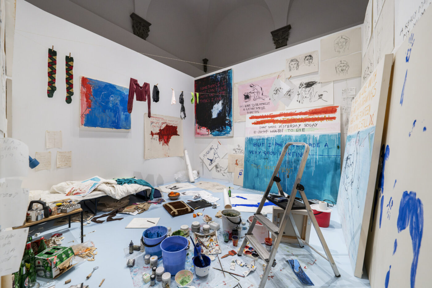 Artistic Haven - Art studio with scattered paint supplies, a ladder, and canvases. Walls display colorful paintings, sketches, and writing. Clothes hang on a makeshift line, and a sleeping area is visible in the cluttered creative space.
