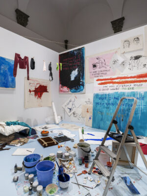 Artistic Haven - Art studio with scattered paint supplies, a ladder, and canvases. Walls display colorful paintings, sketches, and writing. Clothes hang on a makeshift line, and a sleeping area is visible in the cluttered creative space.
