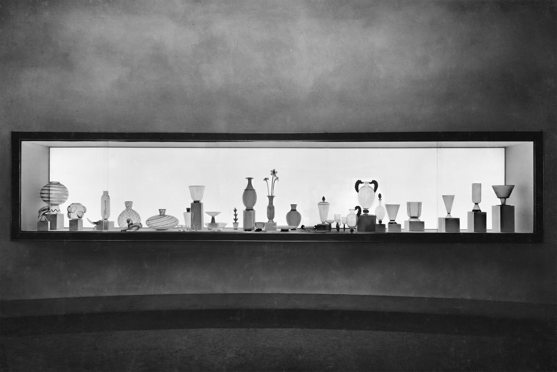 Display of Elegance - A long, rectangular display case showcases various glass objects, including vases, goblets, and abstract forms, arranged in rows against a softly lit background. The wall is a smooth, muted tone, emphasizing the elegant silhouettes of the glassware.