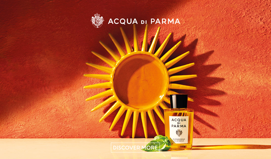 Presentazione di Acqua di Parma" - Una bottiglia di profumo Acqua di Parma è posta di fronte a una parete arancione con una decorazione a raggiera. Un nastro verde è posizionato accanto alla bottiglia. Le parole "Acqua di Parma" e "Scopri di più" appaiono in testo bianco.