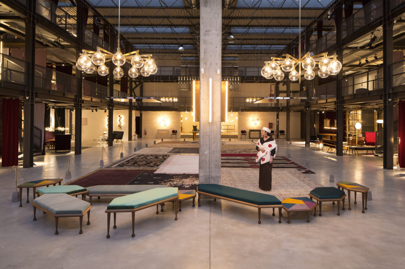 Nina Yashar at Nifular Gallery - Spacious modern atrium with high ceiling, featuring stylish chandeliers, eclectic seating, and various art pieces. The area includes a mix of rugs, benches with multicolored cushions, and a central sculpture, creating an artistic atmosphere.