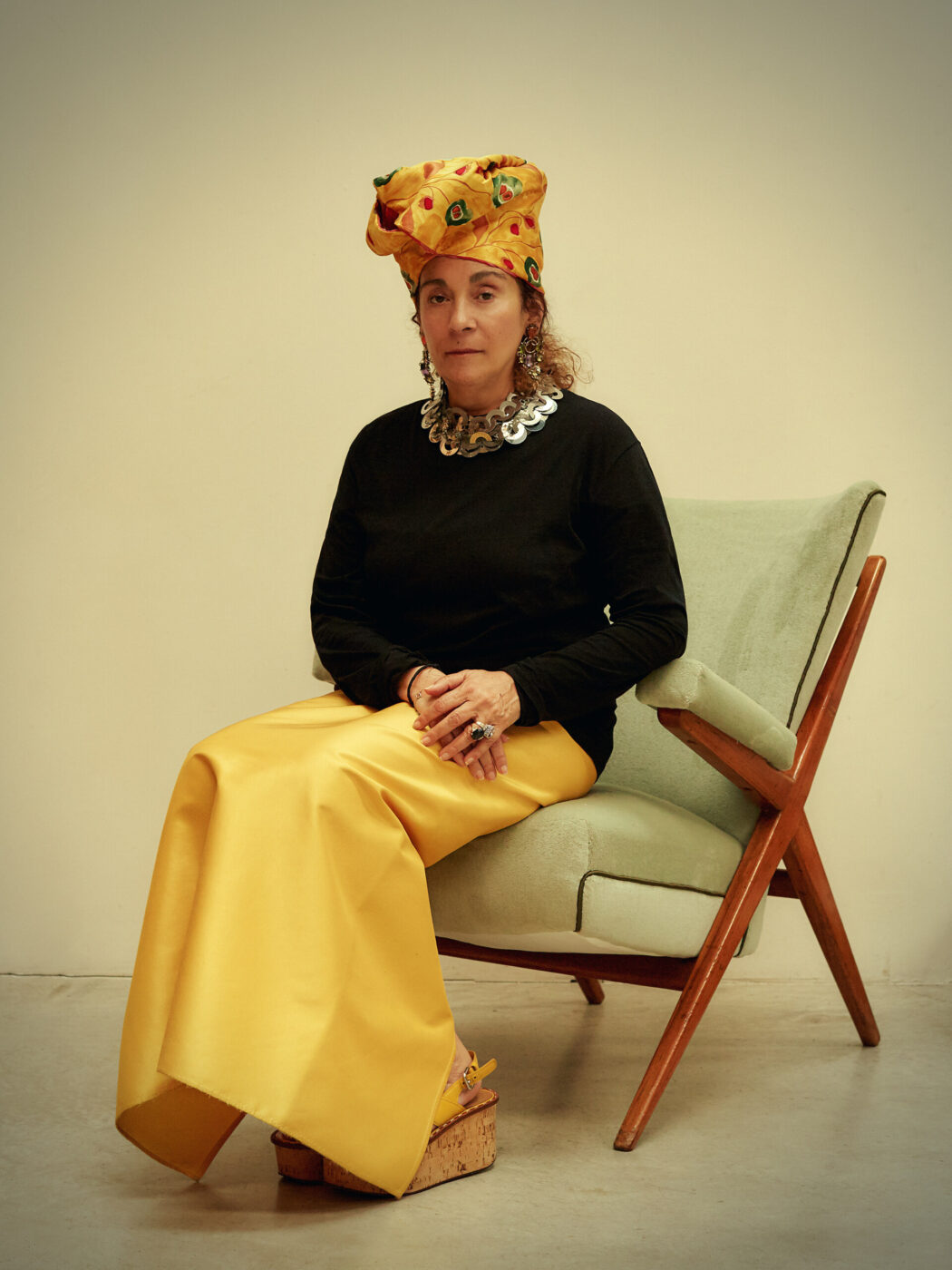 Nina Yashar seated on a retro-style chair, wearing a bright yellow headwrap and skirt, a black top, and a distinctive chunky necklace. They have platform shoes and are gazing calmly at the camera against a plain background. Please provide an image title if you wish to have one included.