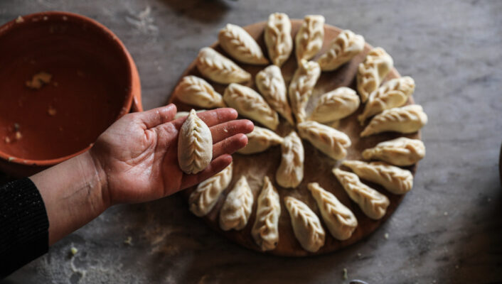 Domu Antiga - A person holds a freshly made dumpling with intricate folds over a wooden board displaying a circular arrangement of similar dumplings. A small bowl and rolling pin are nearby on a gray surface.