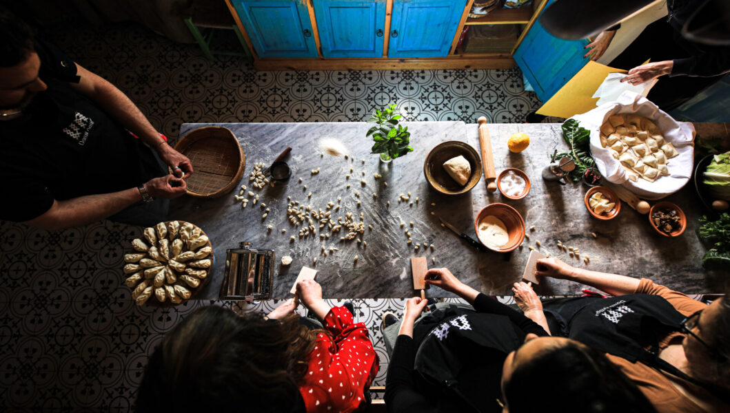 Domu Antiga - Aerial view of a kitchen scene with people making dumplings. Ingredients and kitchenware are scattered on a table. Two people shape dough into dumplings while others assist. Blue cupboards and patterned floor tiles are visible in the background.
