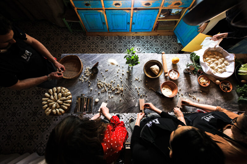 Titolo immagine - Persone riunite attorno a un tavolo che preparano ravioli. Varie ciotole e ingredienti come aglio ed erbe aromatiche sono sparsi sul tavolo. Alcuni individui stanno stendendo l'impasto, mentre altri stanno sistemando i ravioli. Sullo sfondo c'è una credenza con le ante blu.