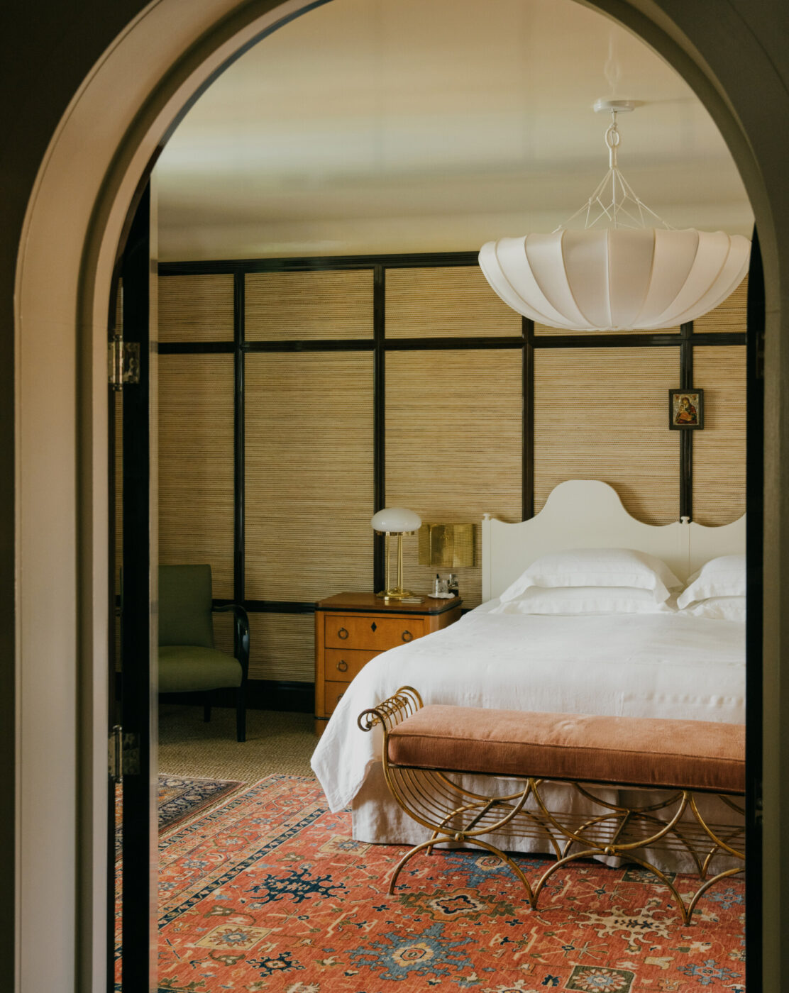Cozy Retreat - Una camera da letto accogliente vista attraverso una porta ad arco, con un letto ben fatto con lenzuola bianche e una panca imbottita. La stanza ha un tappeto a fantasia, un piccolo comodino in legno con una lampada e una poltrona verde. Una lampada a sospensione pende dal soffitto.