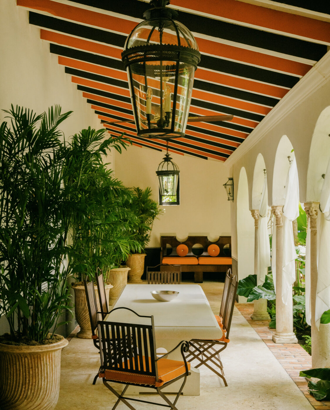 [Titolo dell'immagine] - Un elegante patio in stile mediterraneo con un lungo tavolo da pranzo bianco e sedie in metallo. Lanterne appese e piante in vaso decorano l'area, insieme a colonne ad arco sulla destra. Cuscini arancioni aggiungono un tocco di colore alle sedute.