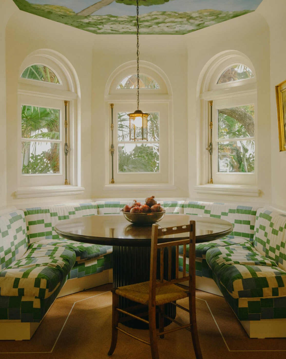 Cozy Breakfast Nook" - Un accogliente angolo per la colazione con un tavolo rotondo in legno circondato da panche imbottite a scacchi bianchi e verdi. Tre finestre ad arco offrono una vista sul giardino. Una ciotola di frutta è appoggiata sul tavolo e una sedia in legno è rivolta verso il tavolo. Una lampada a sospensione è appesa sopra.