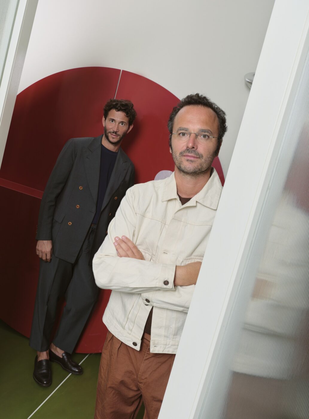 Eligo Studio designers Alberto Nespoli and Domenico Rocca - Two men stand indoors with a red semi-circular wall in the background. The man in front wears glasses, a beige jacket, and has his arms crossed. The man behind is in a dark suit, leaning slightly. The floor is green.