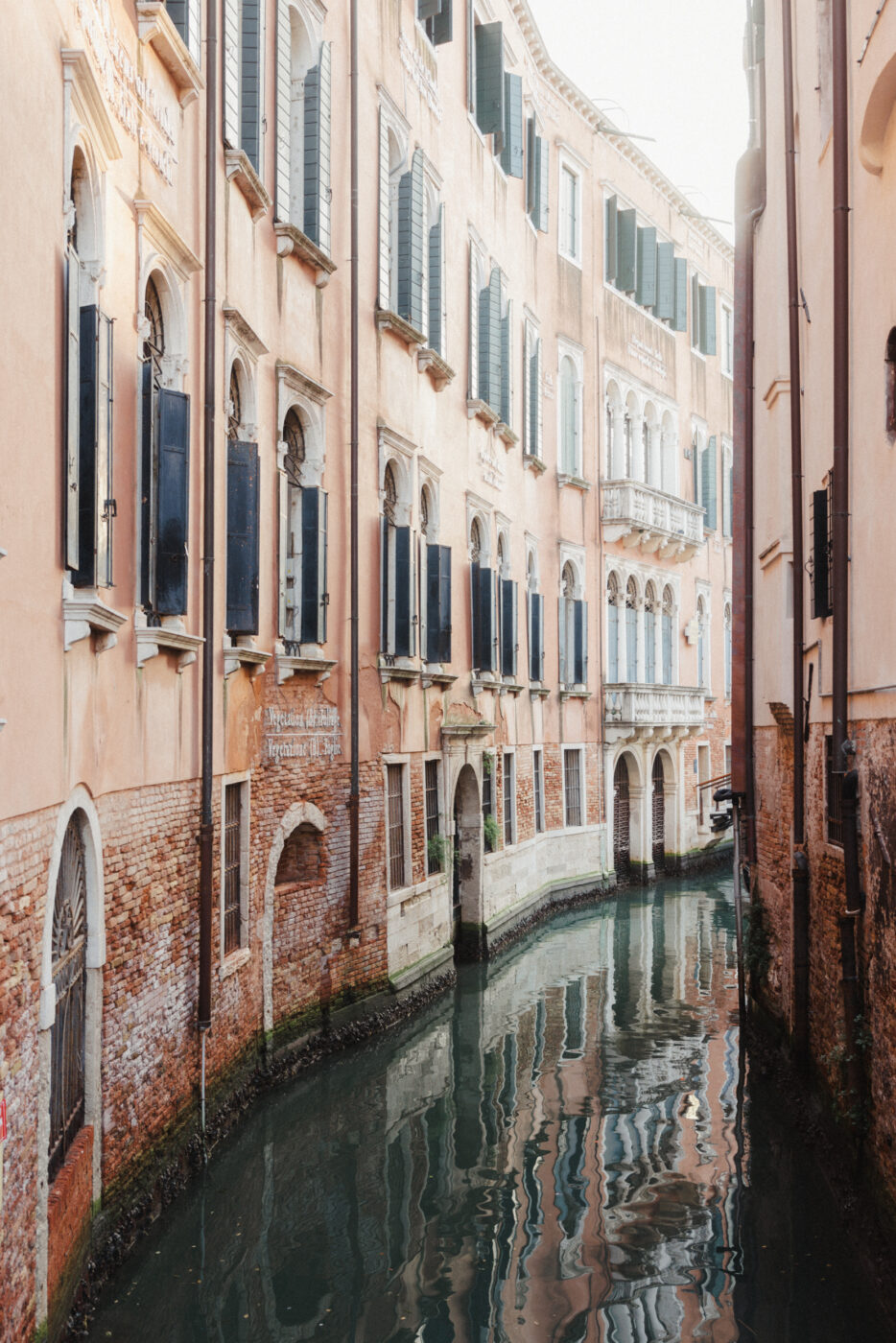 Canale veneziano" - Uno stretto canale a Venezia, fiancheggiato da vecchi edifici invecchiati con finestre ad arco e persiane verdi. La luce del sole pomeridiano crea morbidi riflessi sull'acqua, creando una scena serena e pittoresca.