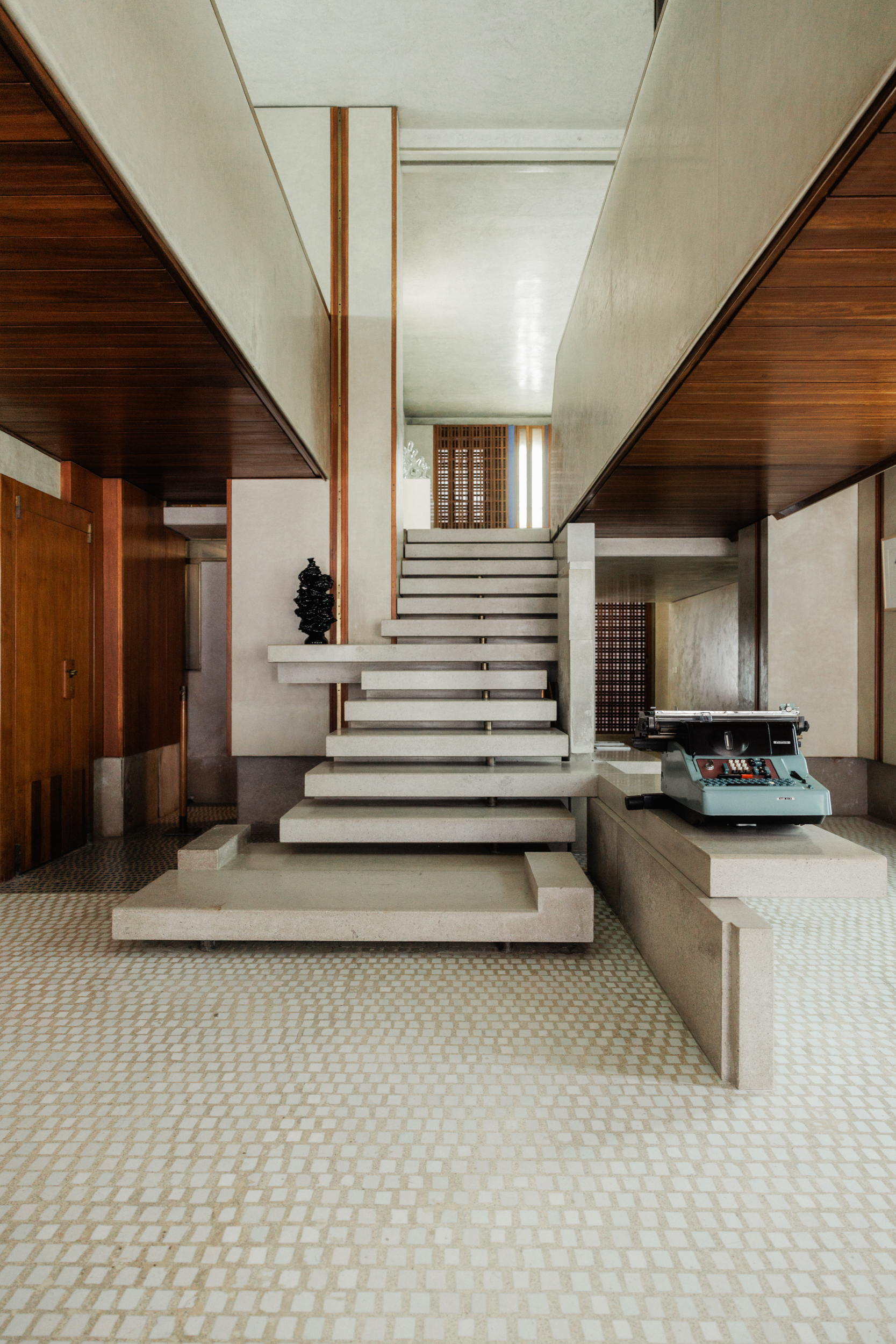 [Interno scala aperta] - Interno con una moderna scala aperta con gradini sospesi e accenti in legno. Una macchina da scrivere è appoggiata su una superficie in pietra sulla destra e il pavimento ha un motivo a mosaico. Lo spazio ha un design minimalista, moderno di metà secolo.