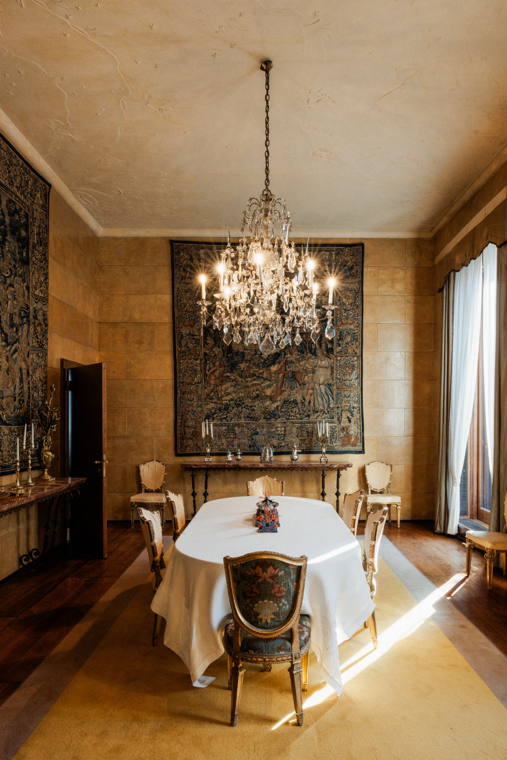 Elegante sala da pranzo - Elegante sala da pranzo con un lungo tavolo coperto da un telo bianco, circondato da sedie ornate. Un grande lampadario è appeso in alto e un arazzo adorna la parete. La luce naturale filtra attraverso alte finestre con tende.