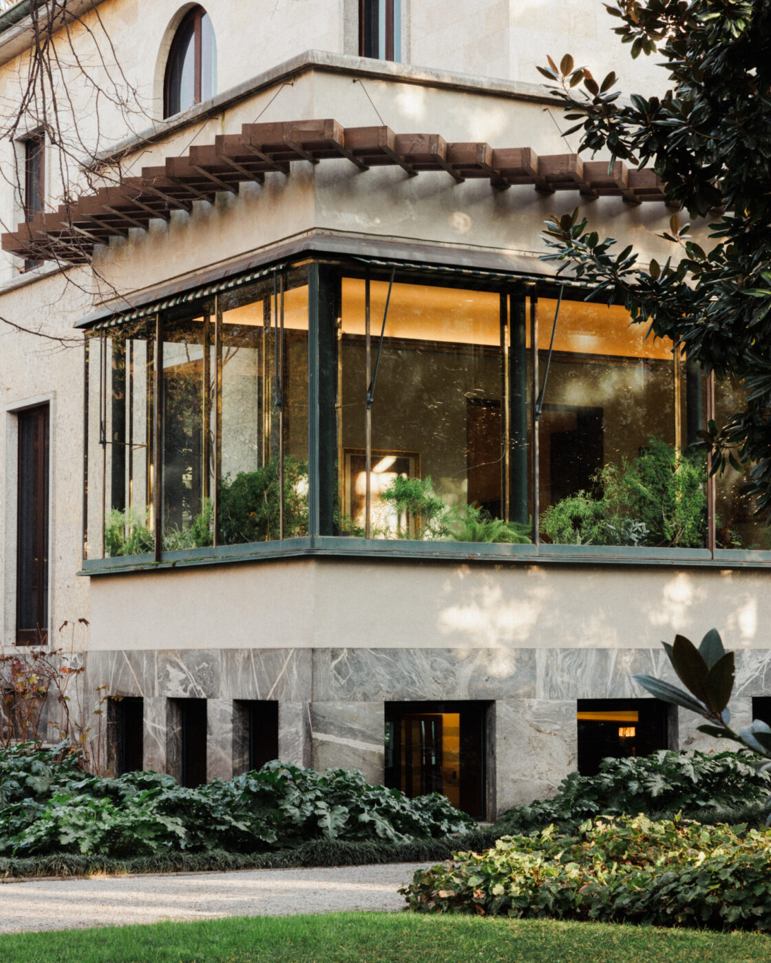 Architettura moderna - Un edificio moderno presenta un angolo con grandi vetrate che espongono rigogliose piante da interno. L'esterno è un mix di pietra e mattoni, circondato da cespugli verdi e un sentiero del giardino ben curato, illuminato da una morbida luce naturale.