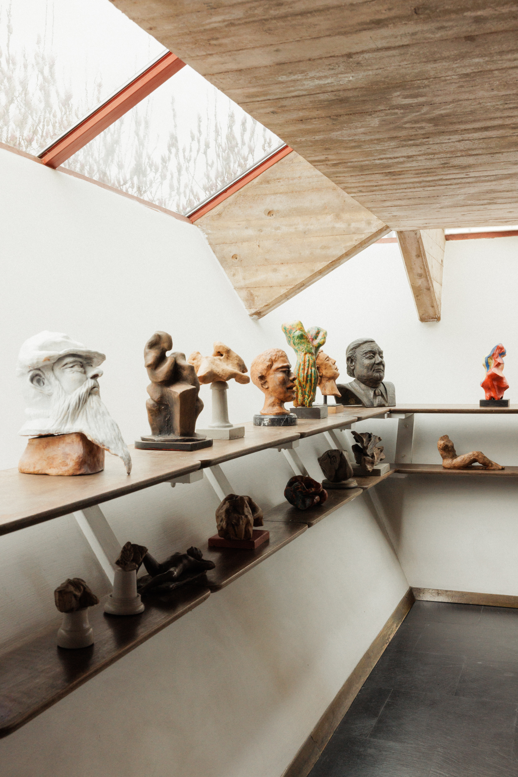 Galleria delle sculture" - Una galleria con varie sculture su mensole di legno sotto un lucernario inclinato. Le sculture includono busti e forme astratte in materiali diversi. Lo spazio ha un design moderno e minimalista con pareti bianche e pavimento in cemento.