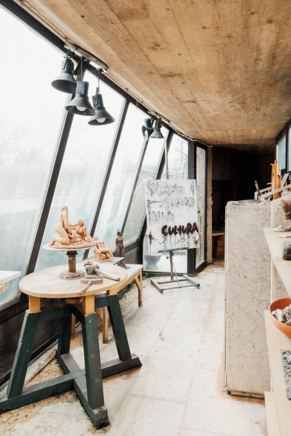 [Titolo dell'immagine] - Uno studio d'arte stretto con finestre inclinate che lasciano entrare la luce naturale. Le sculture sono appoggiate su tavoli di legno e un cavalletto con una tela dipinta è etichettato 'CULTURA'. Vari materiali artistici sono sparsi nello spazio.