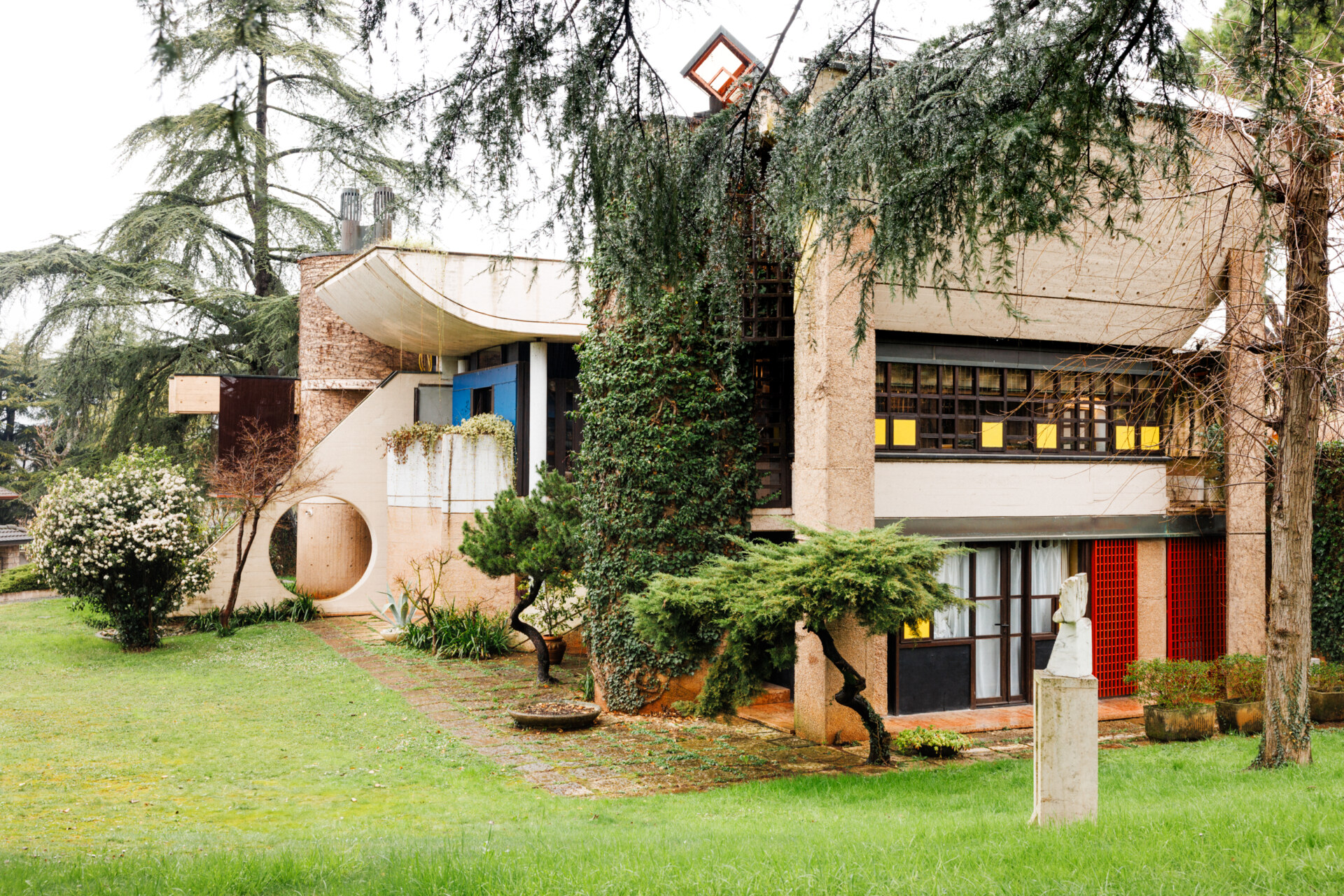 Modernist Haven" - [Alt Text]: Una casa modernista circondata da alberi, caratterizzata da un design unico con forme geometriche. Ha un balcone curvo, accenti rossi e gialli e grandi finestre. Una scultura si erge su un piedistallo nel cortile erboso anteriore.